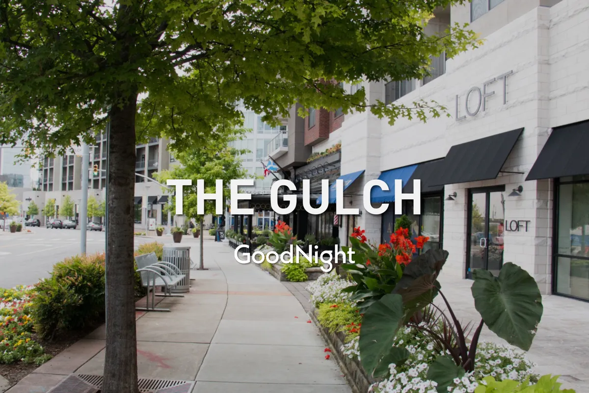 This lively streetscape features tree-shaded sidewalks, flower beds, benches, and storefronts. People are walking, shopping, chatting, and enjoying the welcoming outdoor environment.