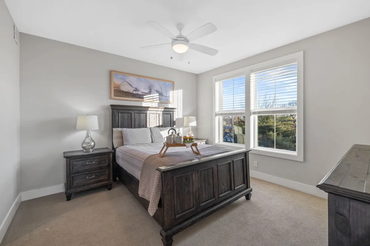 A tranquil bedroom scene featuring a Queen bed with soft bedding, a nightstand with a lamp, windows letting in morning light, and a painting above the headboard. The space feels restful and inviting, ideal for quiet mornings or winding down after a day out.