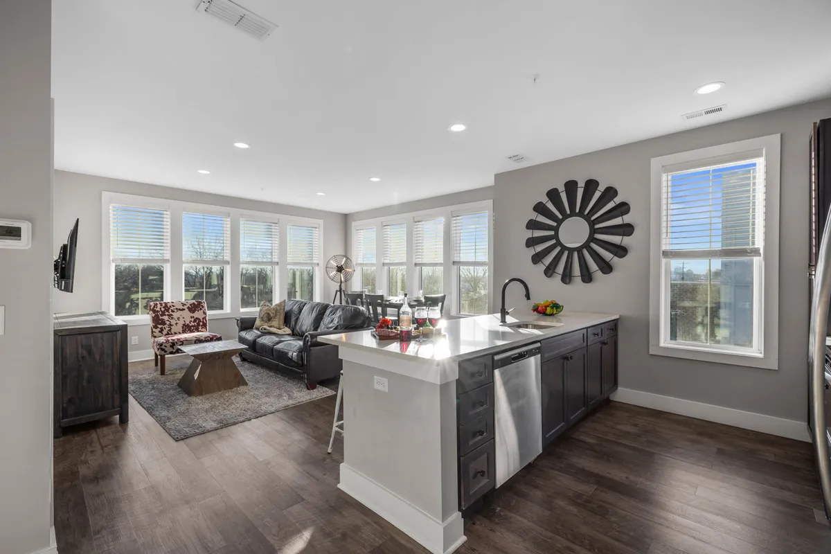The image showcases a bright, open-concept living space with a modern kitchen island, a cozy sofa, large windows flooding the room with natural light, and tasteful decor including a decorative mirror. The scene invites guests to relax, cook, and entertain in a stylish yet comfortable setting.