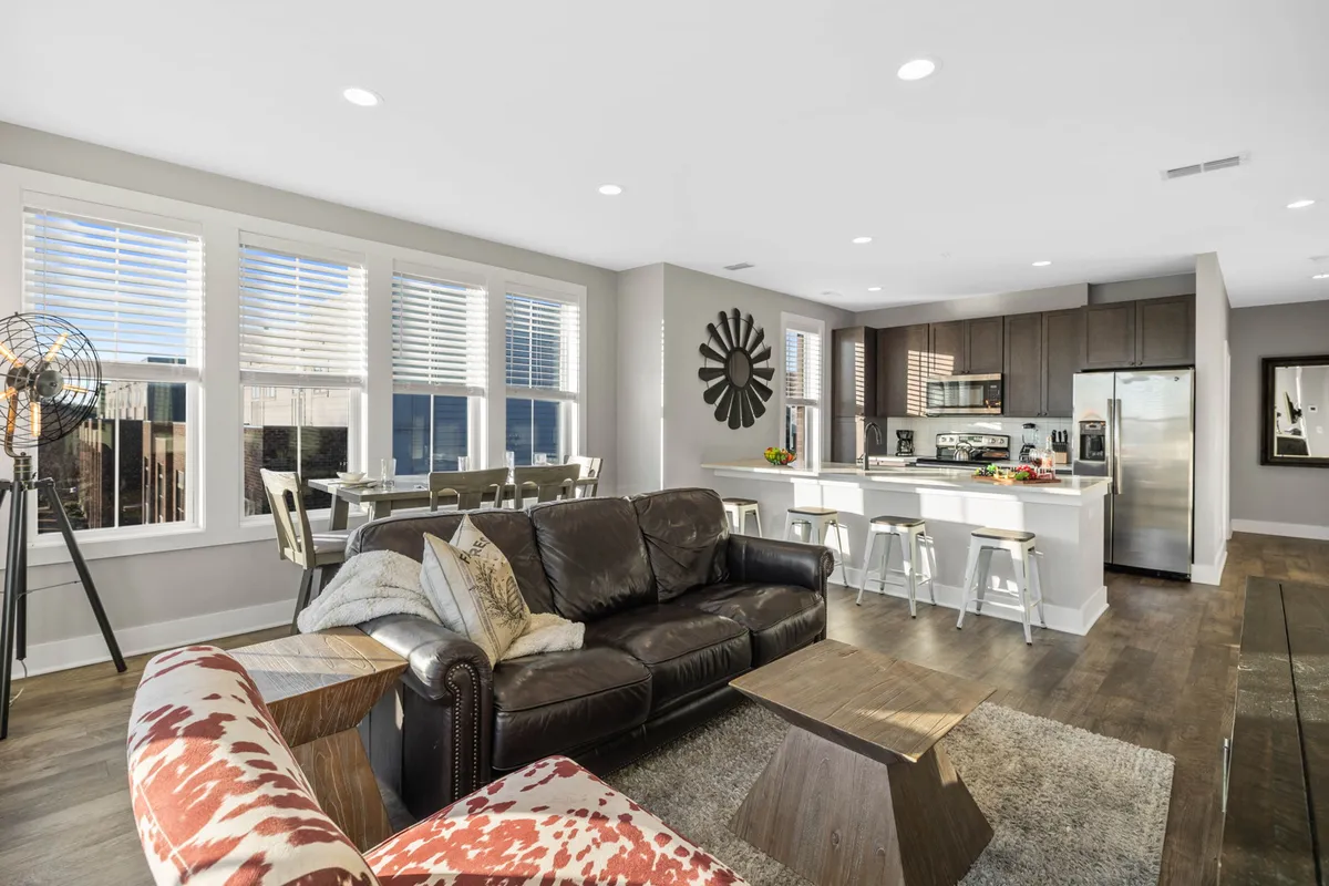 A warm and inviting open-plan space with a leather sofa, a wooden coffee table, a dining table for group meals, a view of the kitchen counter, and decorative accents like a wall clock. The area is designed for family gatherings and entertaining guests, blending living, dining, and kitchen spaces seamlessly.