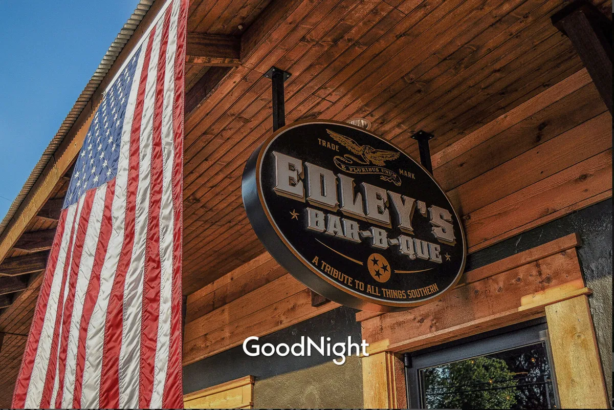 A festive exterior of Edley's Bar-B-Que, complete with an American flag, wooden structure, and signage. The scene suggests a social, music-filled environment with opportunities for live music nights and themed gatherings.